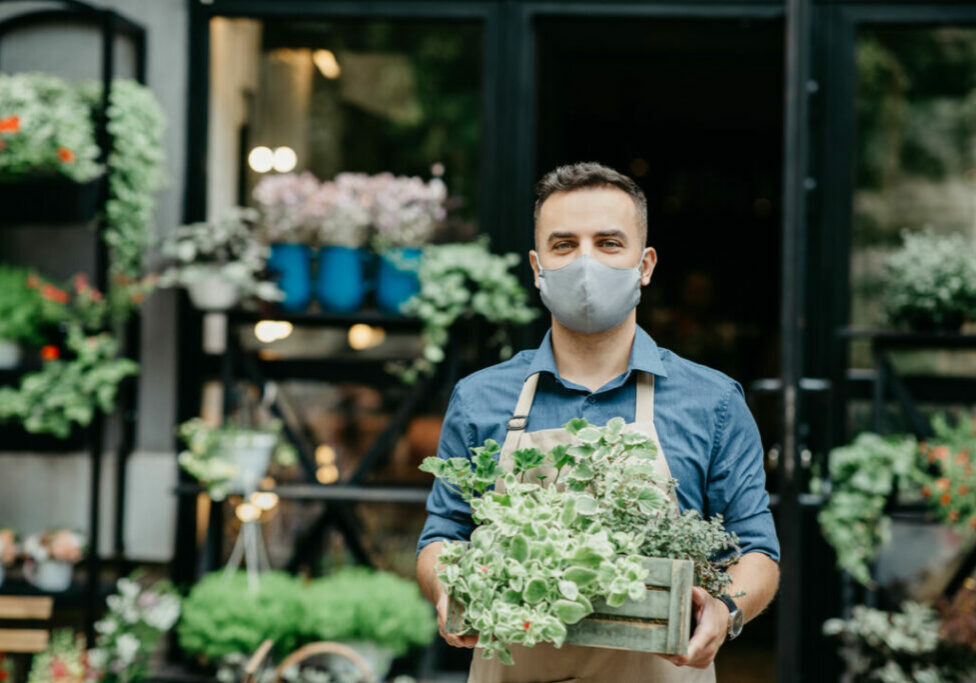 Small business and start of working day. Man in protective mask takes out box of plants outside in front of flower shop